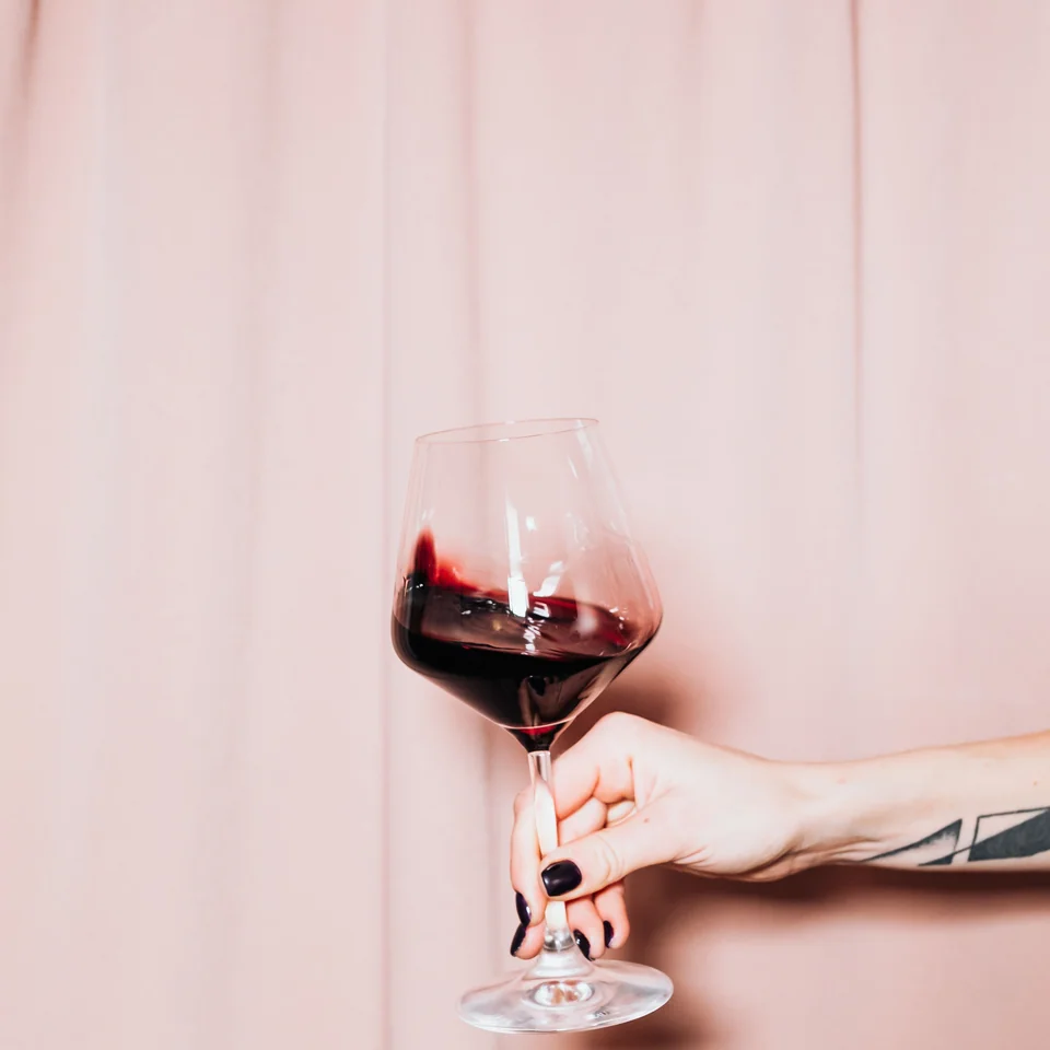 A hand holding a glass of red wine against a pink wall
